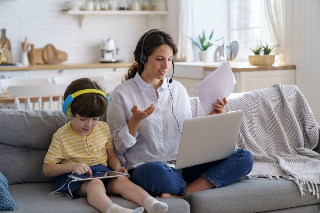 Freelancer mom sitting on sofa at home office during lockdown work on laptop, kid playing at tablet